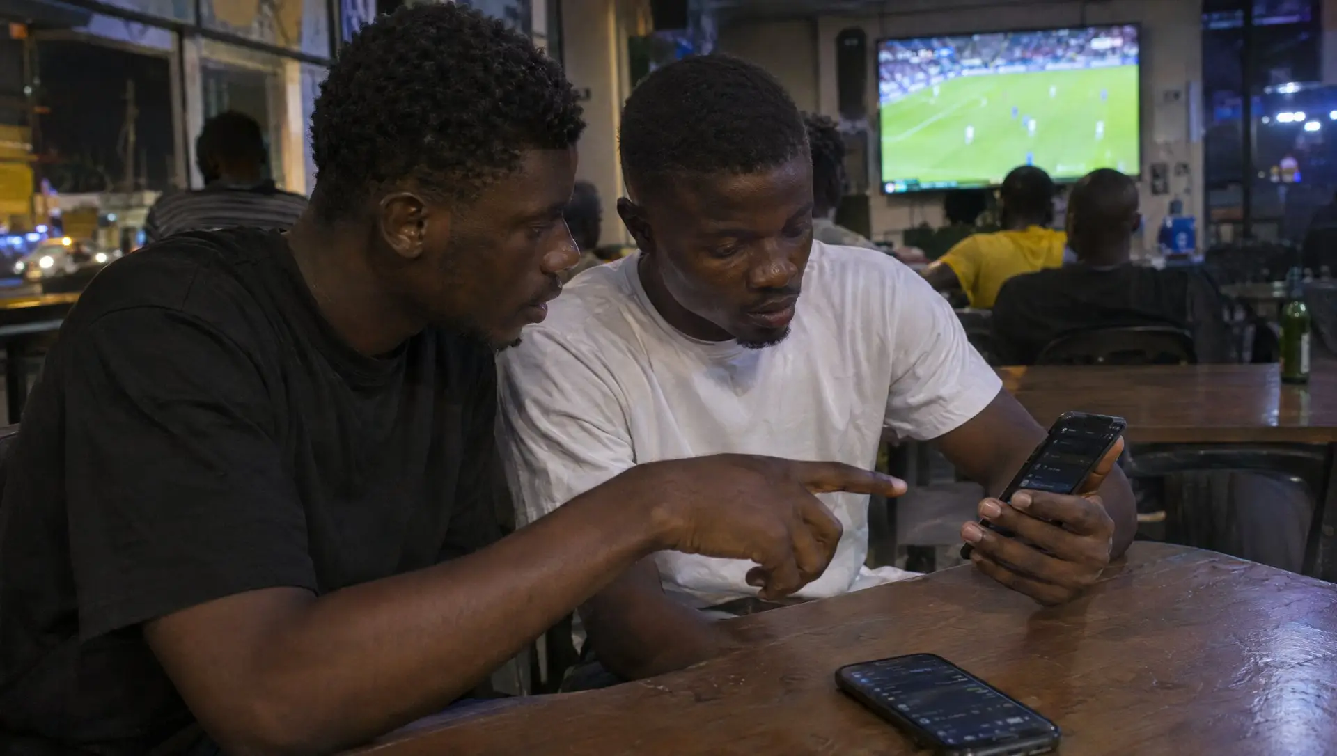 Two Nigerian friends at a Lagos viewing centre checking a bet slip on a phone before a match