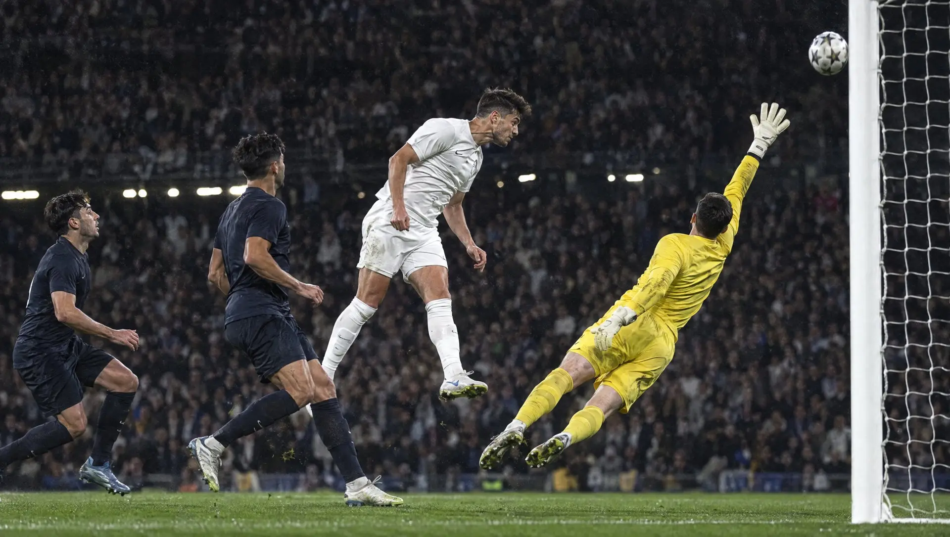 A striker heading the ball past a diving goalkeeper into the top corner under floodlights