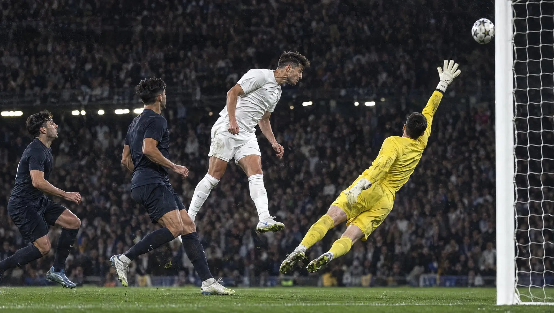 A striker heading the ball past a diving goalkeeper into the top corner under floodlights