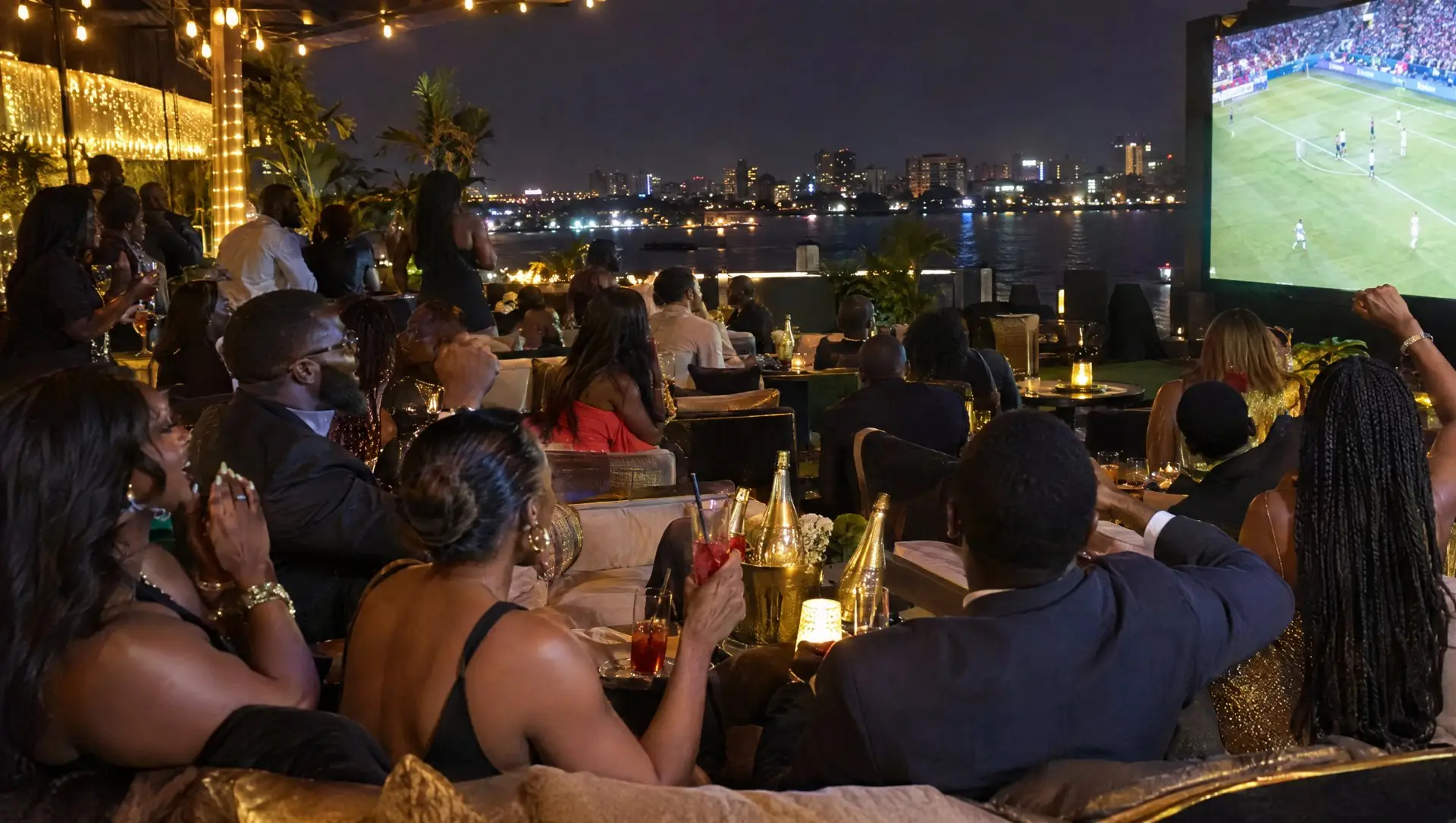 A stylish crowd watching a big European match on a rooftop lounge screen in Victoria Island, Lagos, at night