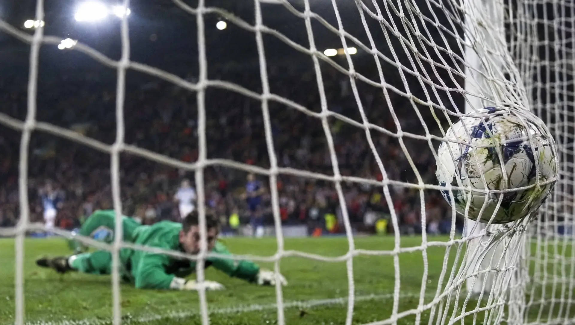 A football hitting the back of the net as the goalkeeper lies beaten on the grass