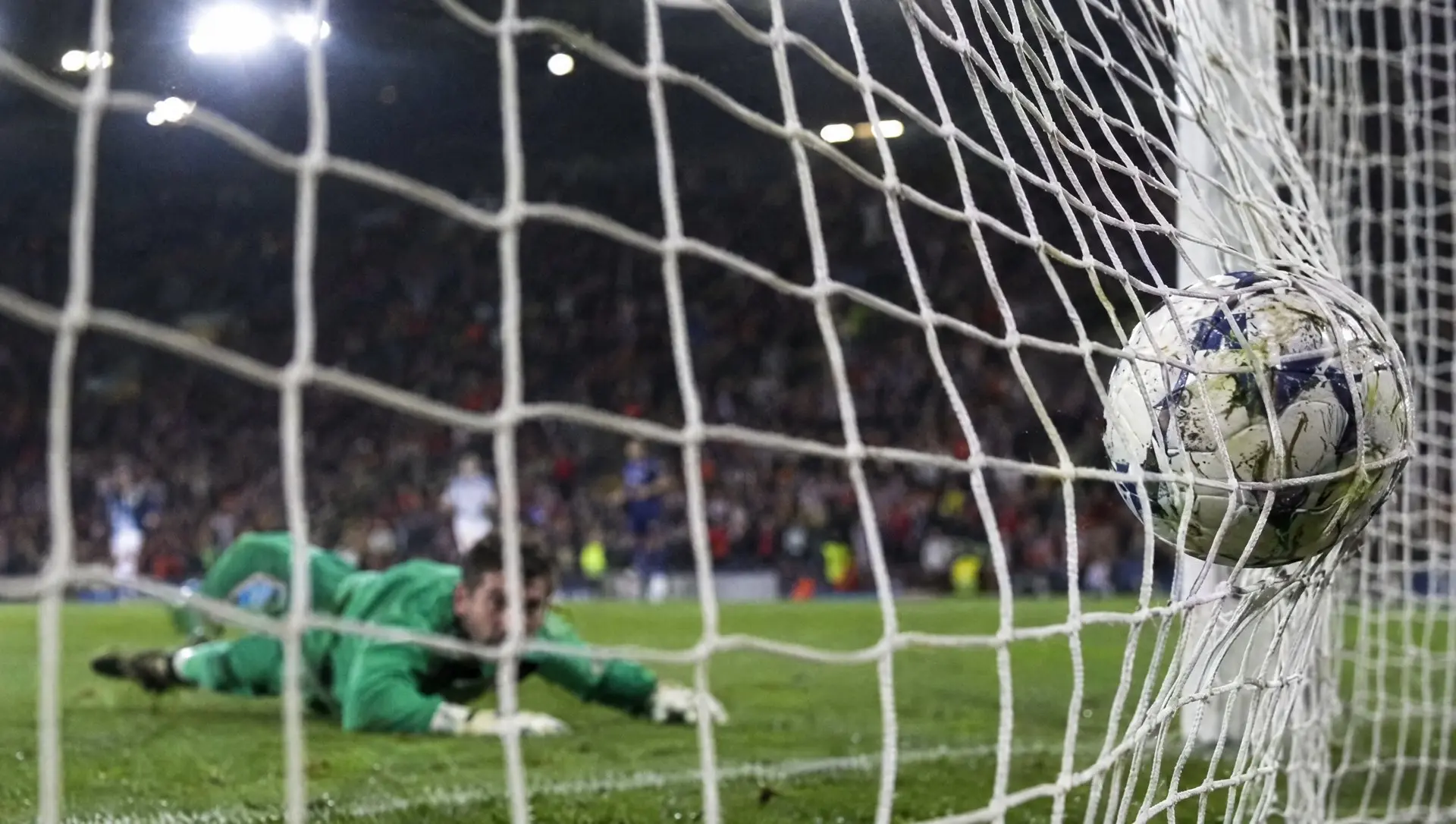 A football hitting the back of the net as the goalkeeper lies beaten on the grass