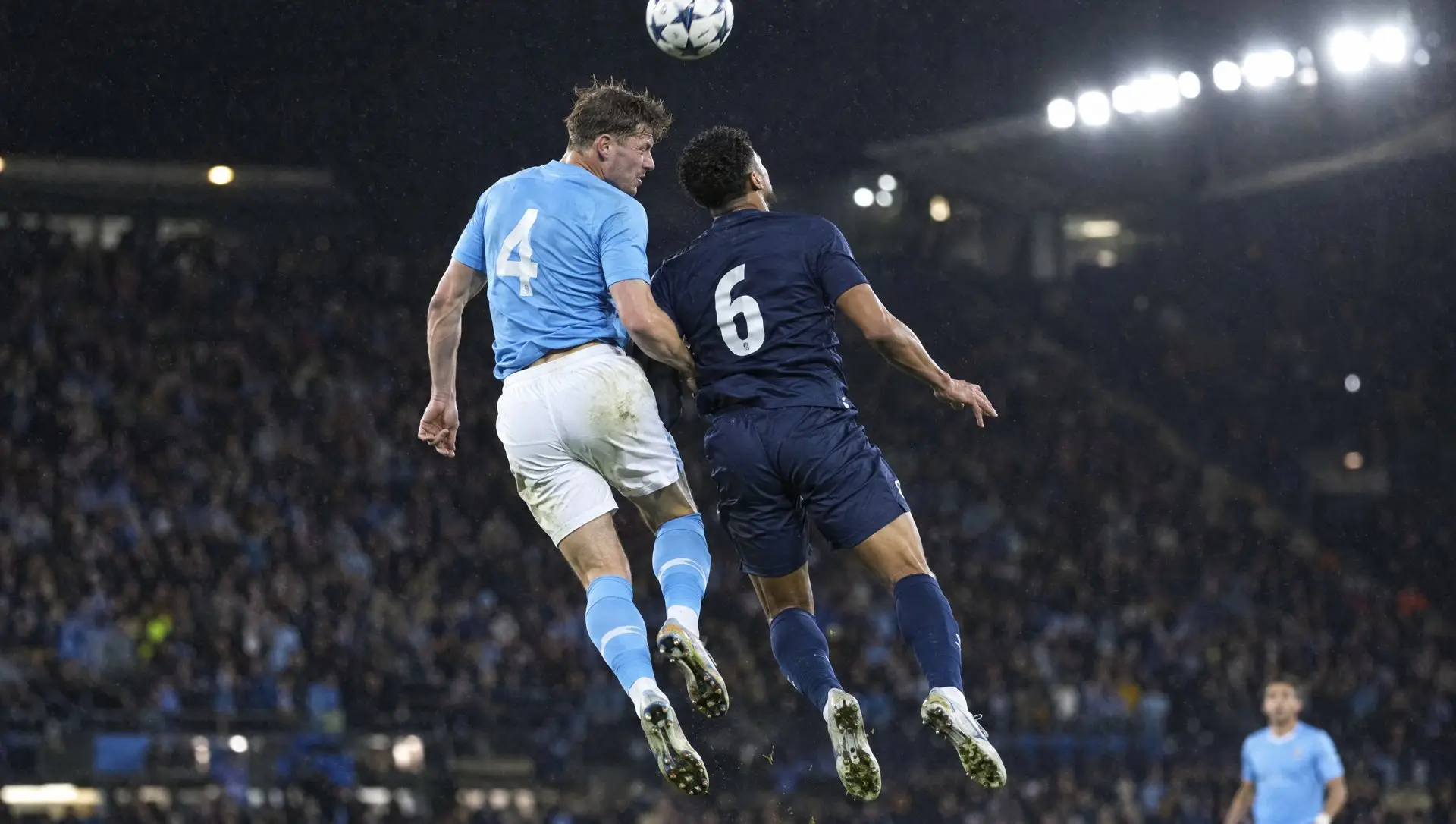 Two footballers in sky blue and dark navy kits challenging for a header under floodlights