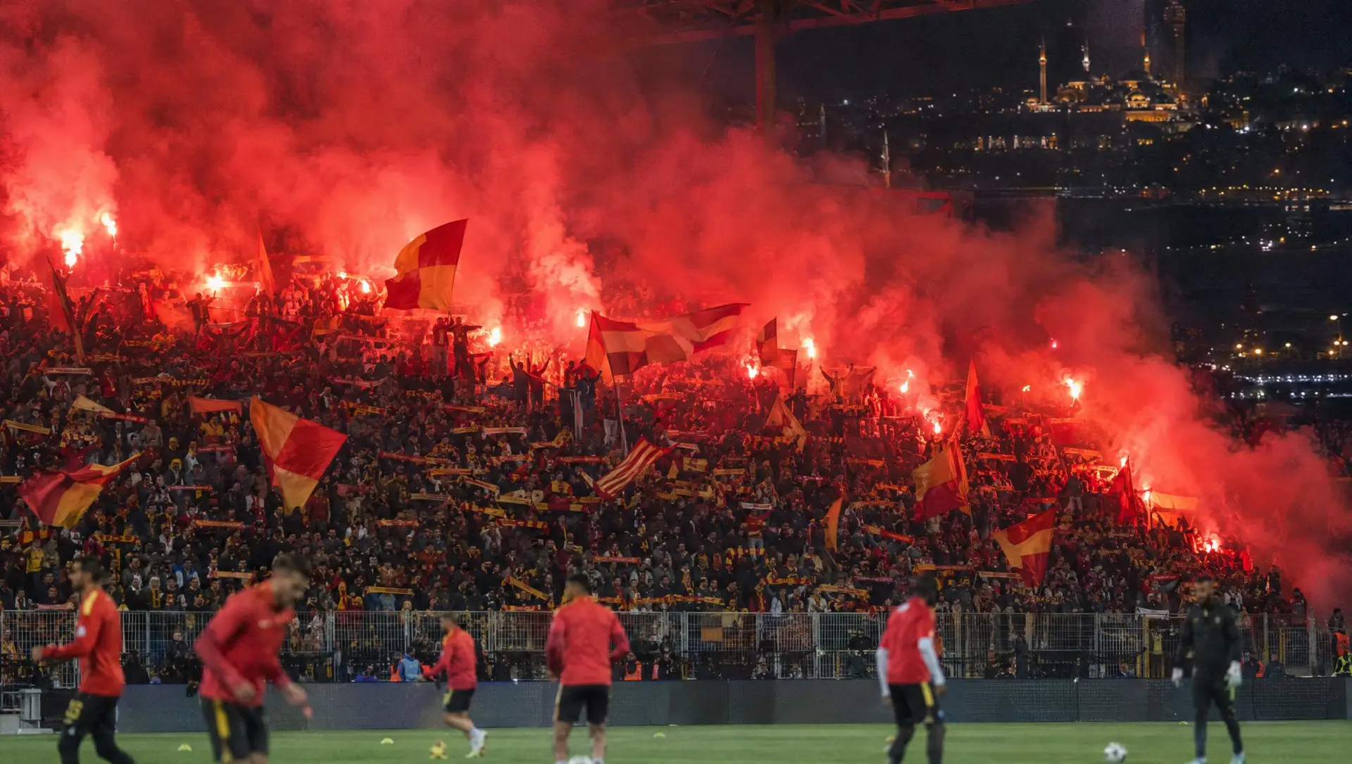 Galatasaray fans in red and yellow creating a wall of noise and flares in an Istanbul stadium at night