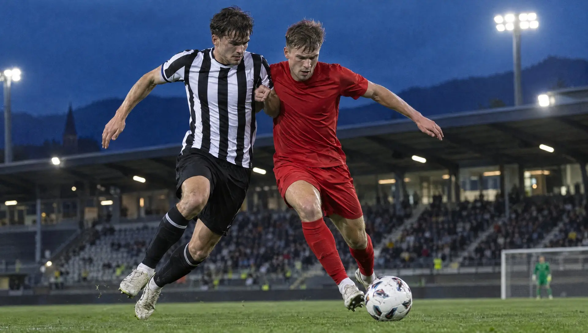 Players in black and white and in red competing for the ball in a compact Austrian stadium at dusk