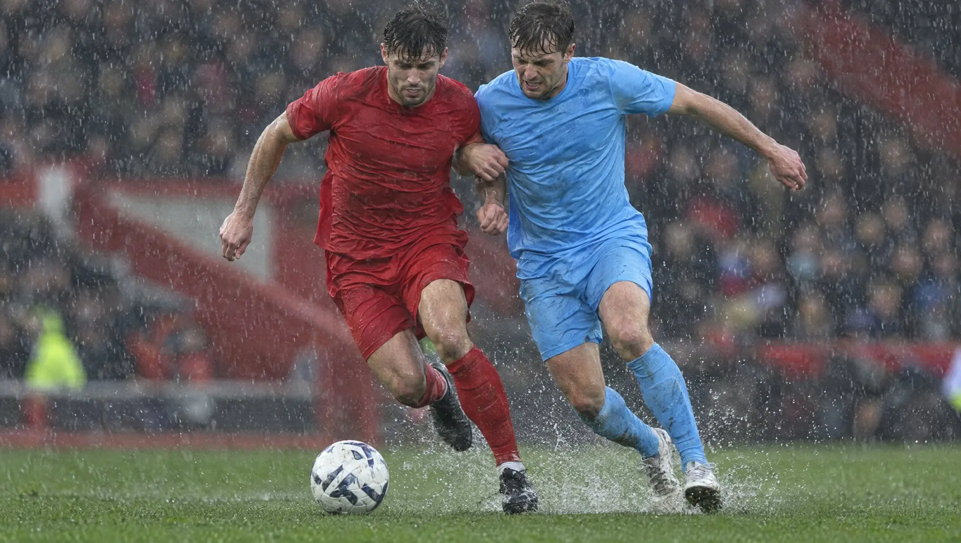 A midfielder in red and a midfielder in sky blue battling for the ball in heavy rain at Anfield-style stadium