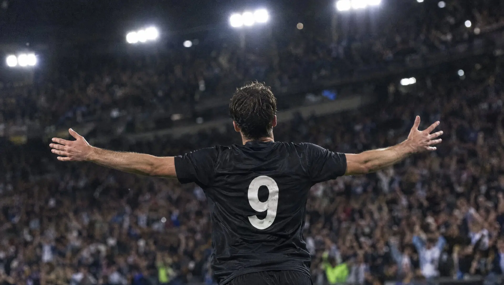 A footballer seen from behind, arms raised to a floodlit crowd after scoring